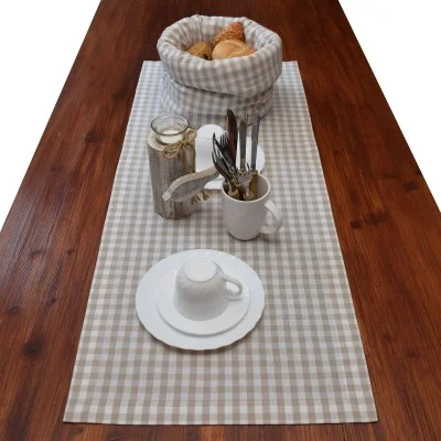 Ein Holztisch mit einem beige-weiß karierten Tischläufer, auf dem ein Brotkorb, ein Glasgefäß, ein Becher mit Besteck und ein weißes Tassen- und Untertassenset stehen.