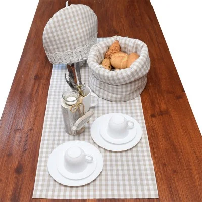 Ein Holztisch mit einem beigefarbenen karierten Tischläufer, zwei weißen Teetassensets, einem silbernen Utensilienhalter und einem passenden Korb mit Deckel für Brötchen und Gebäck.