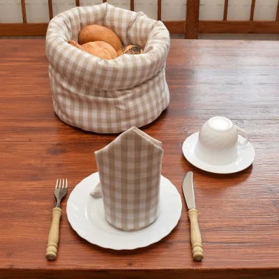 Ein Holztisch ist mit einem weißen Teller, einer Tasse, einer Untertasse, einer Gabel und einem Messer gedeckt. Eine beige karierte Serviette liegt gefaltet auf dem Teller. Ein passender Brotkorb mit verschiedenen Brotsorten steht daneben.
