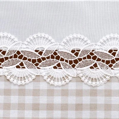 Nahaufnahme eines dekorativen Stoffmusters mit weißer, filigraner Spitzenbordüre auf braunem Grund, oben eingefasst von einfarbigem weißem Stoff und unten von beigefarbenem und weißem Gingham-Stoff in einem Karomuster.
