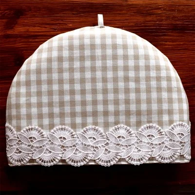 Ein beige-weißer Teewärmer mit Gingham-Muster und abgerundeter Oberseite steht auf einer Holzplatte. Der Teewärmer ist mit einem verschnörkelten Streifen aus weißer Spitze am unteren Rand verziert und hat eine kleine Stoffschleife in der oberen Mitte.