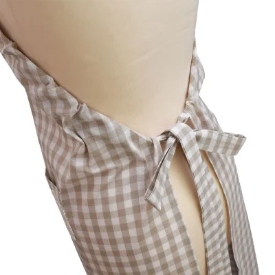 Eine Nahaufnahme einer beige-weiß karierten Schürze, die mit einer Schleife um die Taille einer Schaufensterpuppe gebunden wird. Der Stoff ist in der Taille gerafft, und die Schleife ist in der Mitte hervorgehoben. Der Hintergrund ist neutral und unaufgeregt.