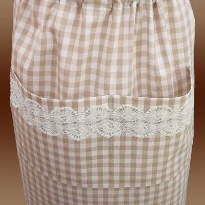 Eine Nahaufnahme einer beige-weiß karierten Schürze mit einer großen Tasche. Die Tasche ist mit einem Streifen weißer Spitze verziert, die ein kompliziertes Blumenmuster aufweist. Der Hintergrund ist ein brauner Farbverlauf.