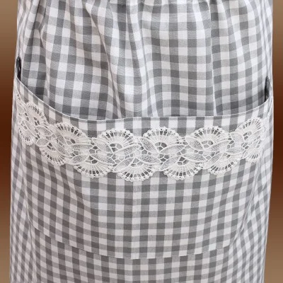 Eine Nahaufnahme einer grau-weiß karierten Schürze mit einer breiten Tasche. Die Tasche ist mit einer dekorativen weißen Spitzenborte mit einem komplizierten floralen und geometrischen Muster versehen. Der Hintergrund ist sanft in Brauntönen verschwommen.