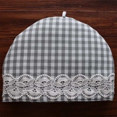 Ein grau-weiß karierter Teewärmer steht auf einer Holzfläche. Der Teewärmer ist kuppelförmig, hat oben eine weiße Schleife und ist am unteren Rand mit einem Band aus verschlungener weißer Spitze verziert.