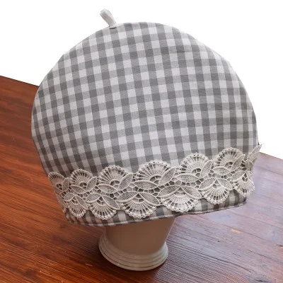 Ein runder grau-weiß karierter Teewärmer mit einer Schlaufe an der Oberseite steht auf einem Holztisch. Der Teewärmer hat ein dekoratives Band aus cremefarbener Spitze am unteren Rand. Er bedeckt eine Teekanne, die größtenteils verdeckt ist.