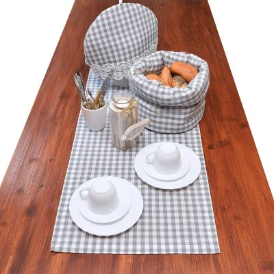 Ein Holztisch mit einem grau-weiß karierten Läufer, einem passenden Brotkorb, einem gedeckten Teller, zwei weißen Tassen mit Untertassen, einer silbernen Teekanne und einer Tasse mit Besteck. In dem Korb liegen Brötchen.