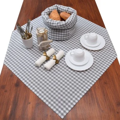 Ein Holztisch mit einem grau-weiß karierten Tischtuch, zwei weißen Tassen und Untertassen, gerollten Servietten mit goldenen Ringen, einem Brotkorb, einem Glas und einer Tasse mit Besteck.