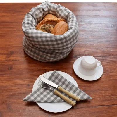 Ein mit grau kariertem Stoff ausgelegter Korb enthält eine Auswahl an Brot. Daneben stehen eine weiße Tasse mit Untertasse und ein Teller mit Serviette, Gabel und Messer auf einem Holztisch.