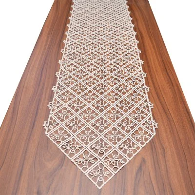 Ein beiger Spitzentischläufer mit geometrischem Blumenmuster ist in der Mitte eines langen, braunen Holztisches platziert. Der Läufer hat spitze Enden und ein kompliziertes, symmetrisches Ausschnittmuster, das sich optisch von der glatten Holzoberfläche darunter abhebt.