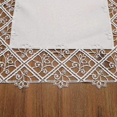 Auf einem Holztisch liegt ein weißes Tischtuch mit einer Spitzenbordüre, die ein geometrisches Muster aus sich kreuzenden Linien und kleinen floralen Mustern zeigt. Der Spitzenrand ist mit detaillierten Blumen und Blättern verziert und zeugt von feiner Handwerkskunst.