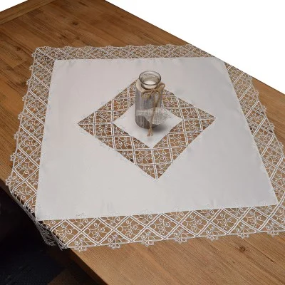 Ein quadratischer Tisch aus hellem Holz ist mit einer weißen Tischdecke bedeckt, die in der Mitte mit einem rautenförmigen Spitzenausschnitt versehen ist. Auf einer sauber gefalteten weißen Tischdecke steht ein mit Schnur verschnürter kleiner Glaskrug.