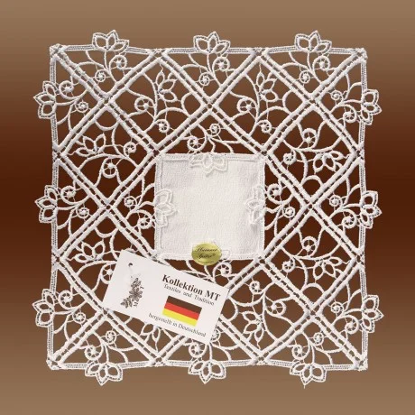 Ein quadratisches, weißes Spitzendeckchen mit einem komplizierten Blumenmuster und einem festen Stoff in der Mitte ist auf einem braunen Hintergrund abgebildet. An dem Deckchen sind ein Schild mit der Aufschrift "Kollektion.MT Textiles and Tradition" und ein goldener Aufkleber mit der Aufschrift "Handmade" angebracht.