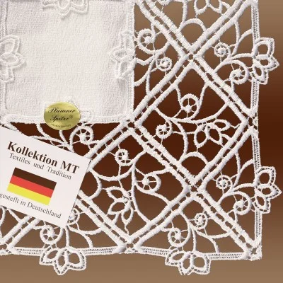 Nahaufnahme eines weißen Spitzenstoffs mit komplizierten Blumenmustern. Eine Karte mit der Aufschrift "Kollektion.MT, Textiles und Tradition, Hergestellt in Deutschland" mit einer deutschen Flagge und einem goldenen ovalen Etikett ist auf dem Stoff abgebildet. Der Hintergrund ist hellbraun.