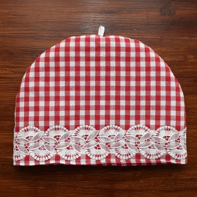 Ein rot-weiß karierter Teewärmer mit geschwungener Spitze steht auf einer dunklen Holzoberfläche. Er hat einen weißen Spitzenbesatz mit Blumenmuster im unteren Bereich und eine kleine weiße Schleife in der oberen Mitte.