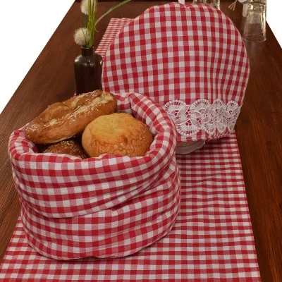 Ein mit rot-weiß-kariertem Stoff ausgelegter Korb enthält verschiedene Brote, darunter eine Brezel und ein Brötchen. Daneben steht eine passende Stoffdecke mit Spitzenbesatz. Beide stehen auf einem Holztisch mit einem passenden Tischläufer, Vasen und Glaswaren im Hintergrund.