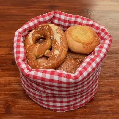 Auf einem Holztisch steht ein rot-weiß-karierter Stoffkorb mit verschiedenen Brotsorten, darunter eine Brezel und runde Brötchen.