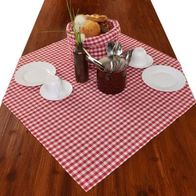 Ein Holztisch mit einer rot-weiß karierten Tischdecke, drei weißen Tellern mit Tassen, einem Brotkorb, einem Behälter mit Utensilien und einer braunen Vase mit Ziergras.