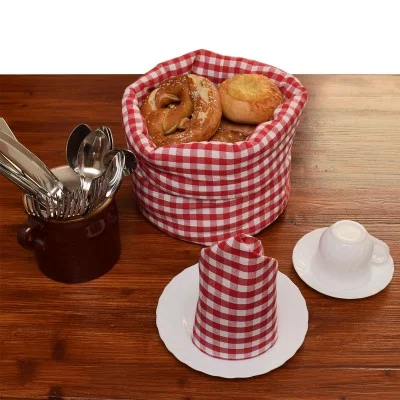 In einem mit rot-weiß-kariertem Tuch ausgelegten Korb liegen Brot und eine Brezel. In der Nähe stehen eine Tasse mit Geschirr, ein gedeckter Teller mit passendem Tuch und eine weiße Tasse mit Untertasse auf einem Holztisch.