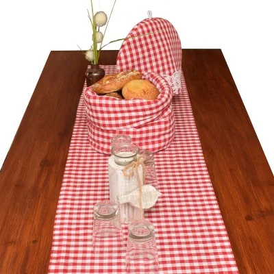 Auf einem Holztisch mit einem rot-weiß karierten Läufer stehen ein passender Brotkorb mit verschiedenen Brötchen, Gläser mit Bändern und Etiketten, Trinkgläser und eine kleine Vase mit weißen Blumen.