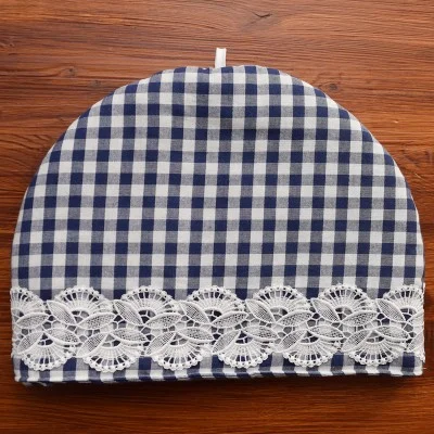 Ein blau-weiß karierter Teewärmer steht auf einer Holzfläche. Er ist halbkreisförmig, hat eine weiße Spitzenborte mit Blumenmuster am unteren Rand und eine kleine weiße Schlaufe am oberen Ende zum Anheben.