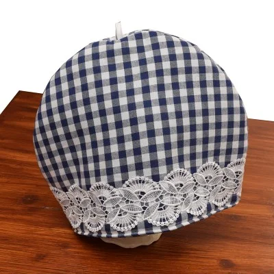 Ein blau-weiß karierter Teewärmer aus Stoff mit einem Band aus weißer Spitze am unteren Rand steht auf einer Holzfläche. Der Teewärmer hat eine runde Form und eine kleine weiße Schlaufe an der Oberseite für eine einfache Handhabung.