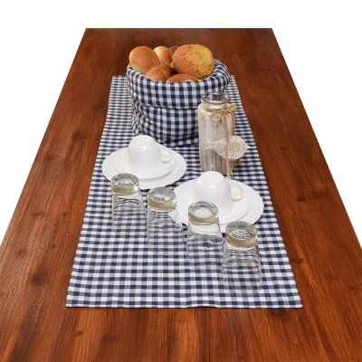 Ein Holztisch mit einem blau-weiß karierten Läufer, auf dem ein Korb mit Brötchen, weiße Tassen und Untertassen, eine Glasflasche mit einer Juteschleife und vier umgedrehte Trinkgläser stehen.