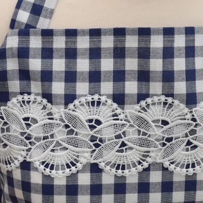 Nahaufnahme eines blau-weiß karierten Stoffes, wahrscheinlich eine Schürze, mit einem horizontalen Streifen aus verschlungener weißer Spitze mit floralen und fächerartigen Mustern, der über den Brustbereich genäht ist.