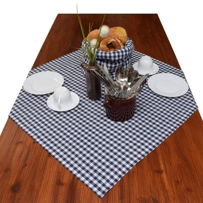 Ein Holztisch mit einem blau-weiß karierten Tischtuch, weißen Tellern, Tassen und Untertassen, einem Brotkorb, einer Vase mit Kunstblumen und einem braunen Behälter mit Utensilien.