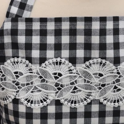 Nahaufnahme eines schwarz-weiß karierten Stoffes, möglicherweise eine Schürze, mit einem breiten horizontalen Band aus verschlungener weißer Spitze mit Blumen- und Blattmotiven über dem Brustbereich. Der Hintergrund ist eine einfarbige, helle Fläche.