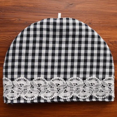 Ein schwarz-weiß karierter Teewärmer mit einer abgerundeten Oberseite, die mit einem weißen Spitzenband in der Nähe des Bodens verziert ist. Der Teewärmer steht auf einer hölzernen Unterlage und hat eine kleine weiße Schlaufe an der Oberseite, um die Handhabung zu erleichtern.