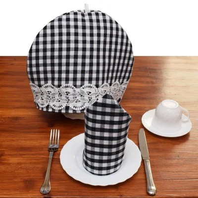 Auf einem gedeckten Tisch auf einer hölzernen Unterlage befinden sich ein schwarz-weiß karierter Teewärmer mit Spitze, eine passende gefaltete Serviette auf einem weißen Teller, eine silberne Gabel und ein silbernes Messer sowie eine weiße Teetasse mit Untertasse, alles fein säuberlich angeordnet.