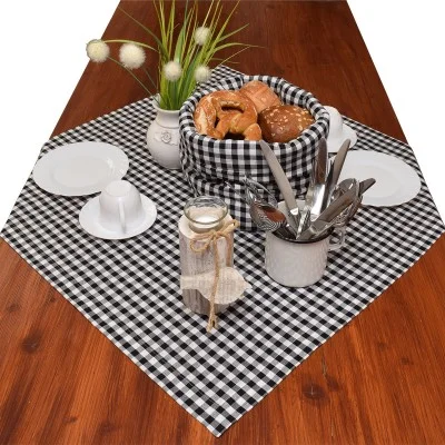 Ein Holztisch mit einer schwarz-weiß karierten Tischdecke, einem Korb mit Brot, Tellern, Tassen, einem mit Schnur umwickelten Glas, einem Becher mit Besteck und einer Vase mit grünem Gras und weißen Blumen.