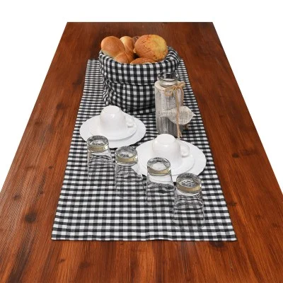 Ein Holztisch, gedeckt mit einem schwarz-weiß karierten Läufer, einem Korb mit Brötchen, drei weißen Tassen mit Untertassen, vier umgedrehten Trinkgläsern und einer Glasflasche mit Korken und Zierschnur.