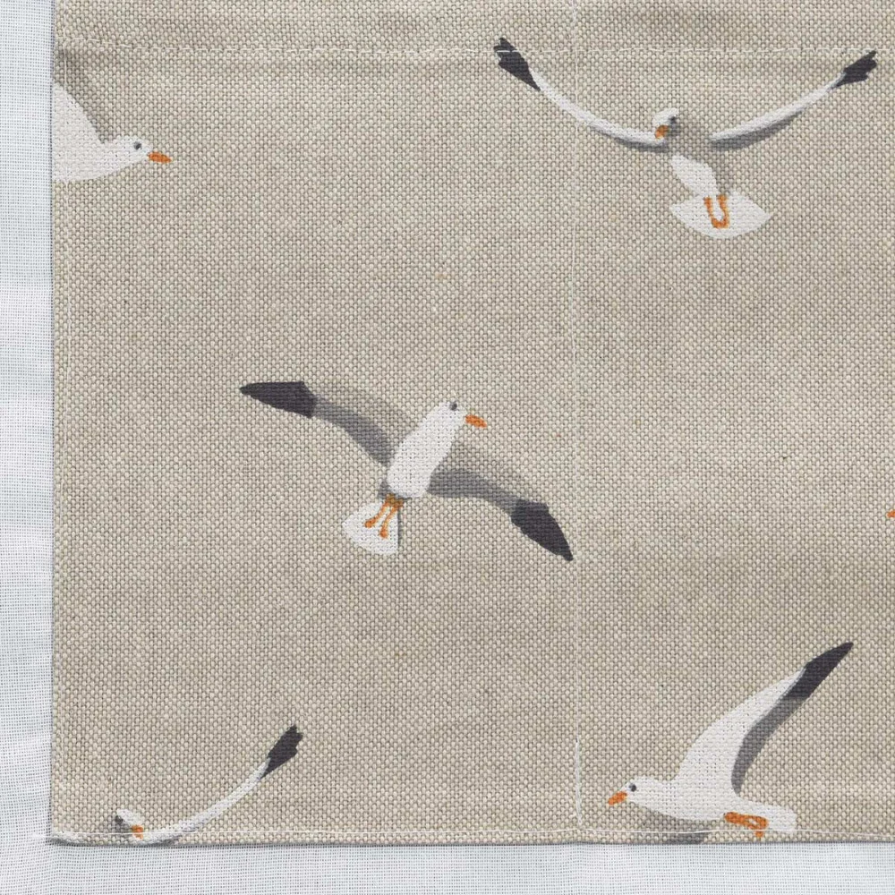 Beigefarbener Stoff mit einem sich wiederholenden Muster aus weißen Möwen in verschiedenen Flugpositionen. Die Möwen haben orangefarbene Schnäbel und Füße mit schwarz bestückten Flügeln. Die Textur des Stoffes ist sichtbar, und am linken Rand befindet sich ein weißer Stoff.