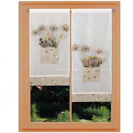 Ein Fenster mit hölzernem Rahmen wird von weißen Gardinen umrahmt. Jedes Vorhangpaneel hat eine beigefarbene Tasche mit aufgestickten Vögeln und Trockenblumen im Inneren. Weiße Spitze ziert die Vorhänge. Draußen sind üppige Grünflächen und ein Gebäude zu sehen.