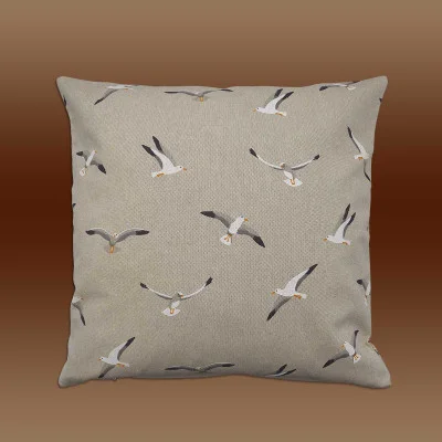 Ein quadratisches, beigefarbenes Kissen ist vor einem Hintergrund mit braunem Farbverlauf platziert. Das Kissen zeigt ein sich wiederholendes Muster aus weißen und grauen Möwen in verschiedenen Flugpositionen, die gleichmäßig über den Stoff verteilt sind. Das Gesamtdesign ist modern.