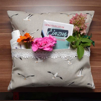Ein beiges Kissen mit Möwenaufdruck und einer Tasche mit Spitzenbesatz auf der Vorderseite enthält einen kleinen Blumenstrauß, ein Fläschchen mit Lotion und ein Buch. Die Gegenstände sind ordentlich auf einer Holzunterlage angeordnet.