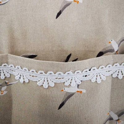 Nahaufnahme eines beigen Stoffes mit Möwenmuster. Auf dem Stoff befindet sich eine Tasche, die von einer weißen Spitzenborte mit Wellenrand eingefasst ist. Die Spitze befindet sich oberhalb der Tasche und hebt sich von dem strukturierten Stoff ab.