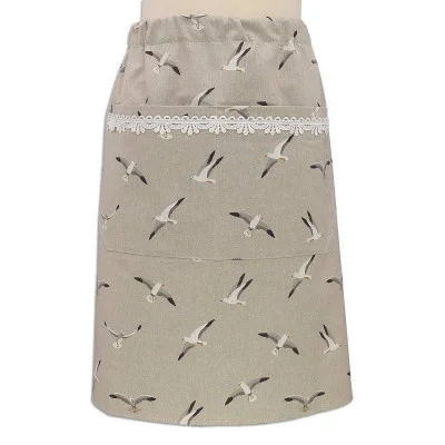 Eine beigefarbene Taillenschürze mit einem Muster aus fliegenden Möwen, einer geräumigen Vordertasche und einem Streifen aus weißer Spitze am oberen Rand der Tasche. Die Schürze wird an einer Schaufensterpuppe vor einem einfarbigen Hintergrund gezeigt.
