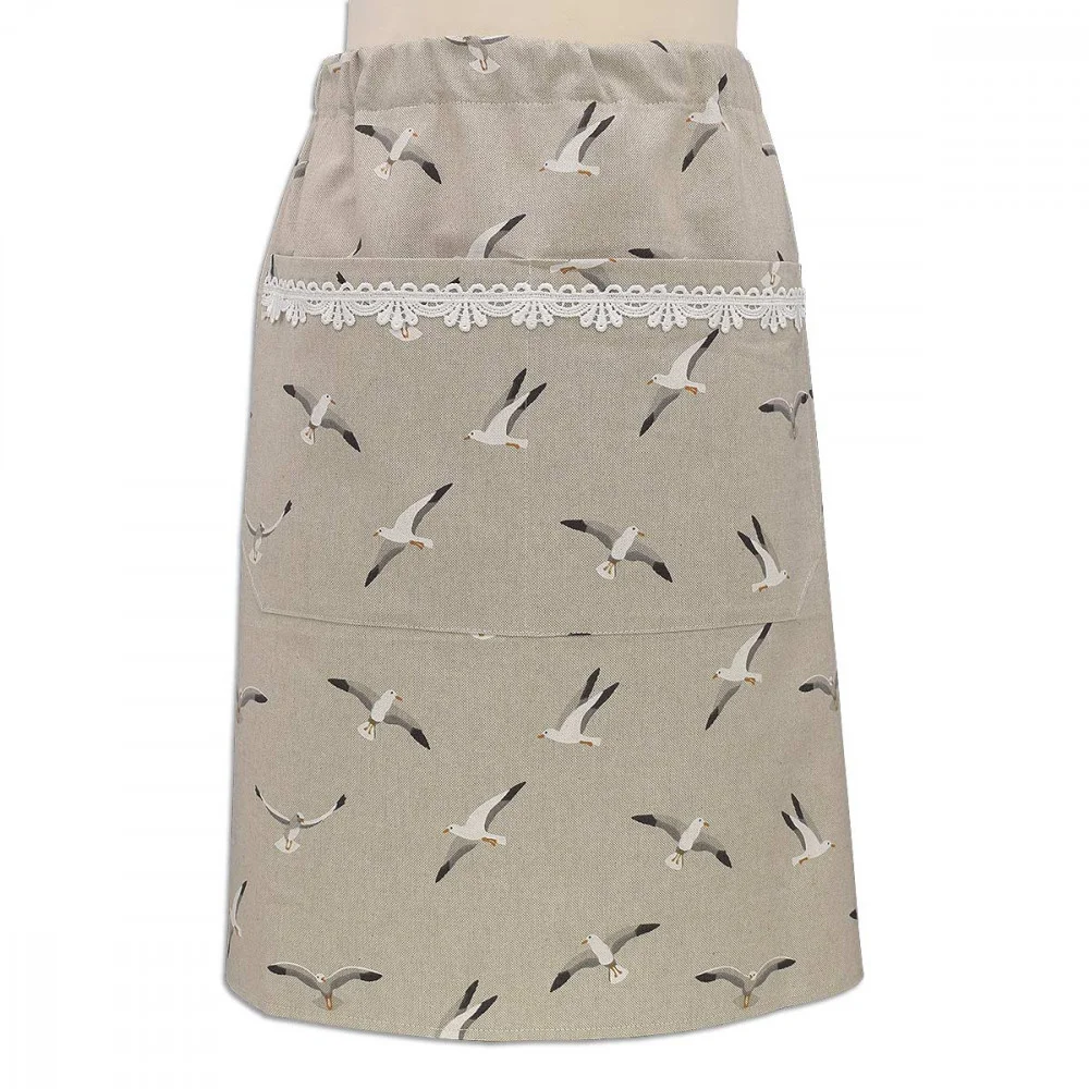 Eine beigefarbene Taillenschürze mit einem Muster aus fliegenden Möwen, einer geräumigen Vordertasche und einem Streifen aus weißer Spitze am oberen Rand der Tasche. Die Schürze wird an einer Schaufensterpuppe vor einem einfarbigen Hintergrund gezeigt.