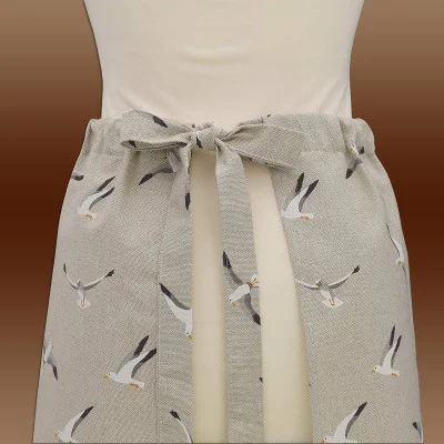 Eine hellbeige Taillenschürze mit einem Muster aus fliegenden Möwen wird im Rücken mit einer Schleife gebunden. Die Schürze bedeckt den unteren Oberkörper und ist an einer Schaufensterpuppe vor einem braunen Hintergrund mit Farbverlauf abgebildet.