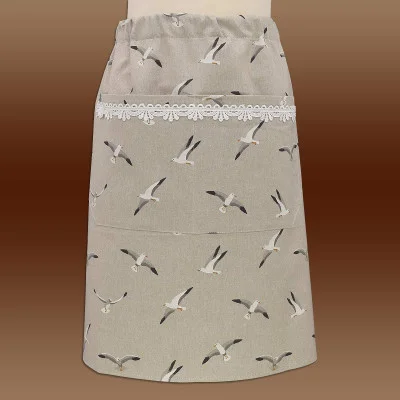 Eine beigefarbene Halbschürze mit elastischem Bund zeigt ein Muster aus fliegenden Möwen. Sie verfügt über eine große Vordertasche mit weißer Spitzenborte. Die Schürze präsentiert sich vor einem braunen Hintergrund mit Farbverlauf.