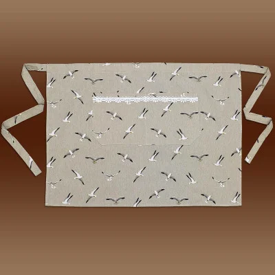 Eine beigefarbene Schürze mit verlängerten Taillenbändern zeigt ein einheitliches Muster aus fliegenden Vögeln. In der Mitte befindet sich eine rechteckige Tasche, die durch einen Streifen weißer Spitzenborte am oberen Rand hervorgehoben wird. Der Hintergrund besteht aus einem braunen Farbverlauf.