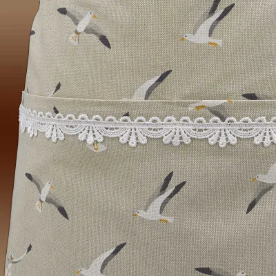 Nahaufnahme eines Stoffes mit einem Muster aus fliegenden Möwen auf einem hellbeigen Hintergrund. Eine weiße Spitzenborte mit einem dekorativen Wellenmuster verläuft horizontal über die Mitte. Der Stoff hat eine sichtbare, strukturierte Oberfläche.