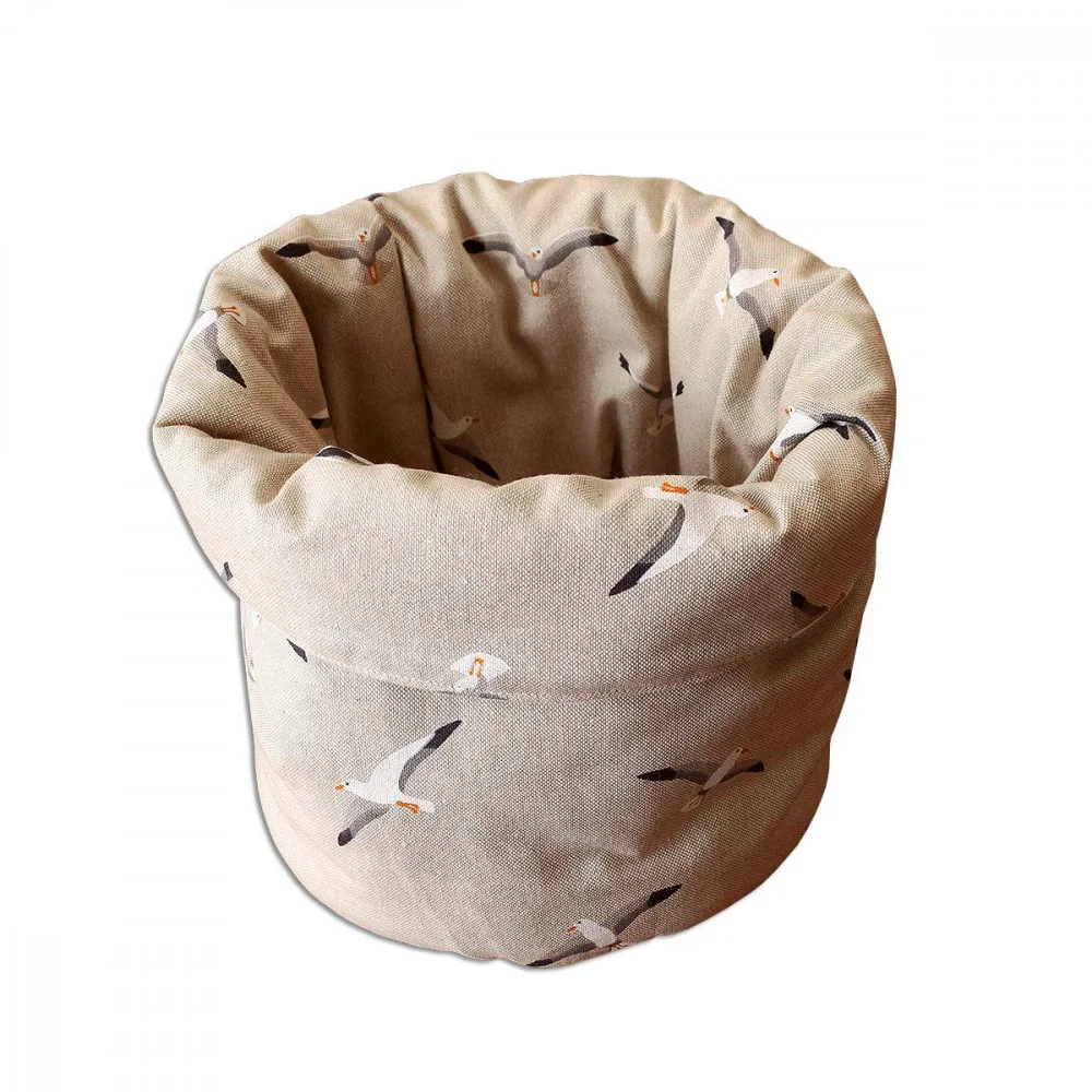 Ein beigefarbener, zylindrischer Stoffkorb mit weichem, gerolltem Rand, auf dem weiße und graue Möwen mit orangefarbenen Schnäbeln und Füßen im Flug auf einfarbigem Hintergrund abgebildet sind. Der Korb ist leer und wird vor einem weißen Hintergrund gezeigt.