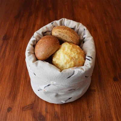 Ein beiger Brotkorb aus Stoff mit einem Vogelmuster enthält drei runde Brötchen - ein einfaches, ein mit Samen bestreutes und ein mit einer knusprigen Außenseite -, die auf einem Holztisch stehen.