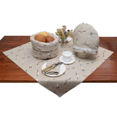 Ein Holztisch ist mit einem beigen Tischtuch mit Vogelmuster bedeckt. Auf dem Tisch stehen ein passender Brotkorb mit Brot, ein Teewärmer, weiße Teetassen und Untertassen, Teller mit Besteck und in der Mitte eine kleine Vase mit Trockenblumen.