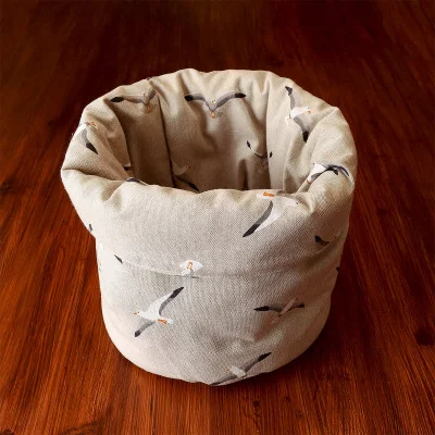 Ein runder, beiger Stoffkorb steht auf einem Holzboden. Der Korb zeigt ein sich wiederholendes Muster von Möwen im Flug, die sich durch weiße Körper, Flügel mit schwarzer Spitze und orangefarbene Schnäbel auszeichnen.