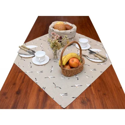 Ein Holztisch mit einer beigen Tischdecke mit Vogelmotiv ist mit weißen Teetassen, Untertassen, Tellern und Servietten gedeckt. Eine Vase mit Trockenblumen, ein Brotkorb mit Brot und ein kleiner Weidenkorb mit Bananen und Äpfeln sind ebenfalls vorhanden.